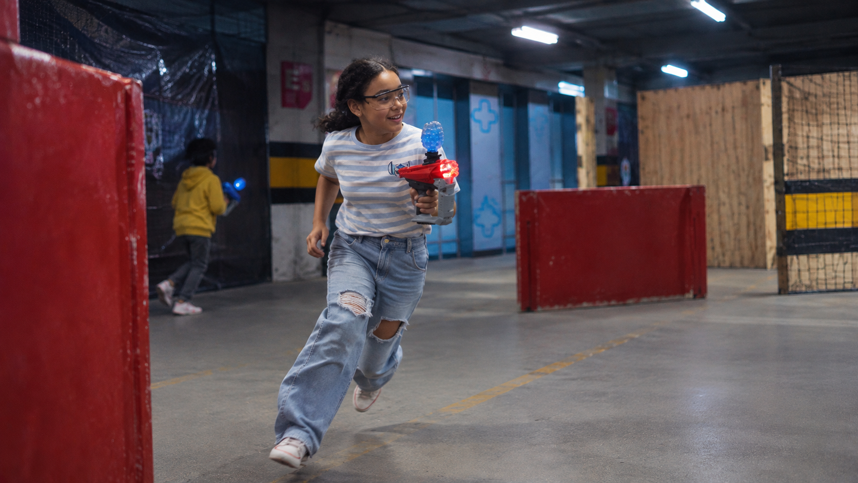 Menina com proteção nos olhos correndo com seu blaster na arena